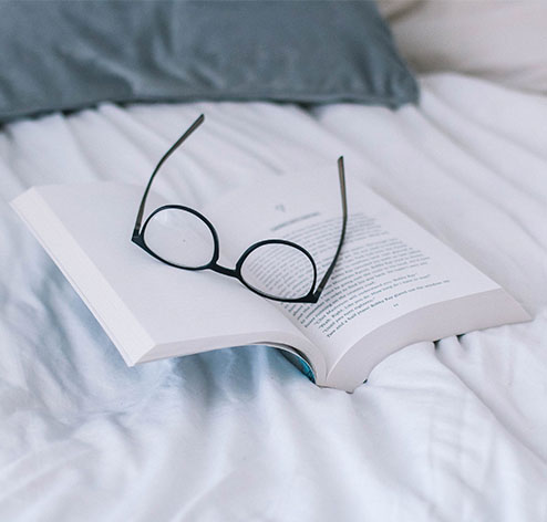 Uma foto que mostra um óculos em cima de um livro aberto em cima de uma cama.